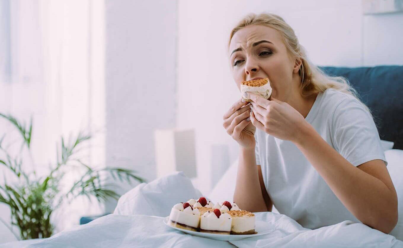 selective focus of sad woman in pajamas eating cake in bed alone